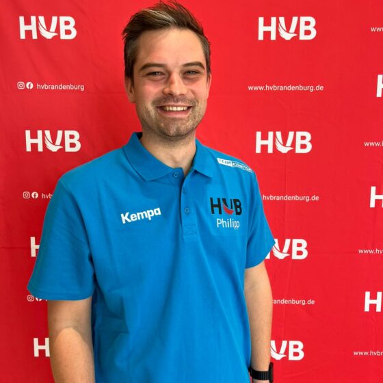 Ein Mann, der ein blaues HVB-Poloshirt mit der Aufschrift „Philipp“ trägt, steht lächelnd vor einem roten Hintergrund, auf dem sich die Logos von HVB und hvbrandenburg.de wiederholen.