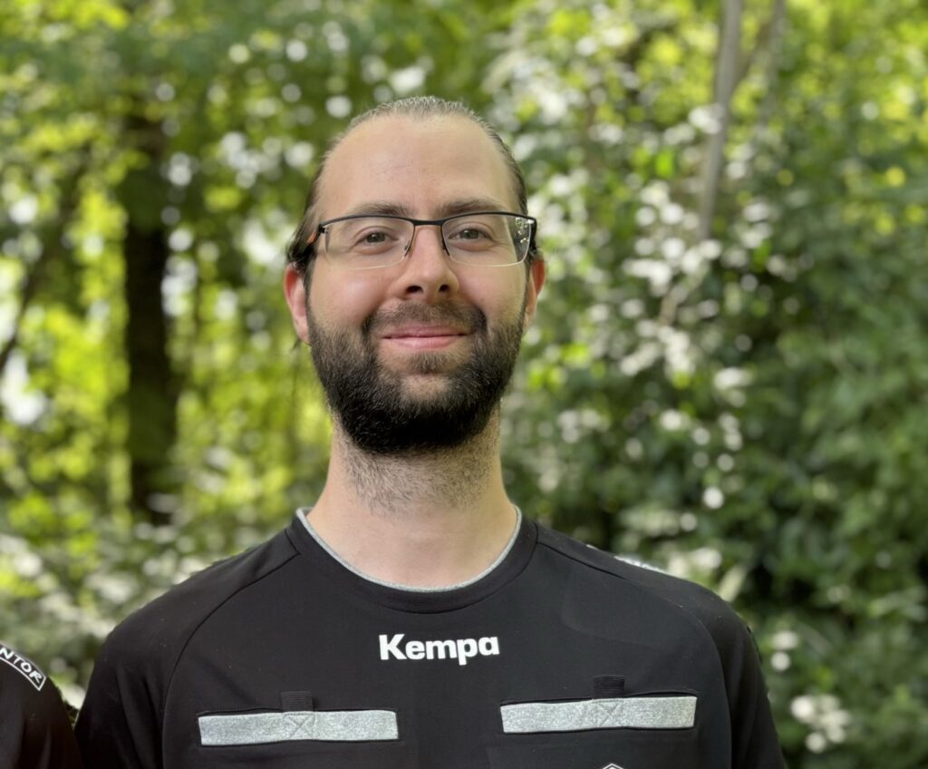 Ein Mann mit Brille und Bart steht im Freien vor grünem Laub und trägt ein schwarzes Kempa-Sporttrikot mit weißen und roten Aufnähern, was sein Engagement für den Nachwuchsleistungssport widerspiegelt.
