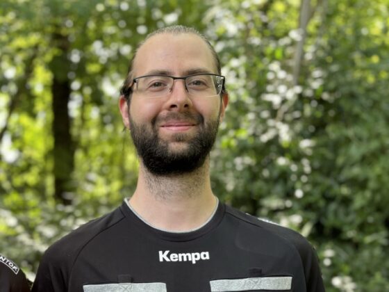 Ein Mann mit Brille und Bart steht im Freien vor grünem Laub und trägt ein schwarzes Kempa-Sporttrikot mit weißen und roten Aufnähern, was sein Engagement für den Nachwuchsleistungssport widerspiegelt.