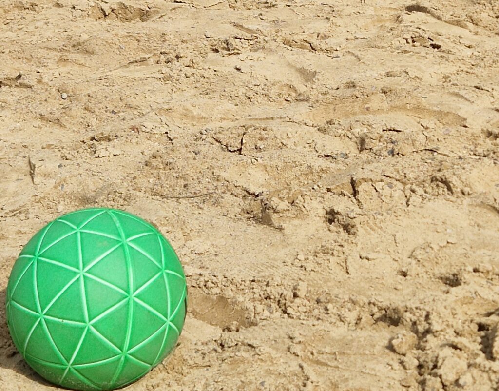 Ein grüner Ball mit geometrischen Mustern liegt im Sonnenlicht auf sandigem Boden und vermittelt die Energie des Nachwuchsleistungssports. Der trockene, strukturierte Sand bedeckt den größten Teil des Bildes und lässt eine lebendige Arena für angehende Sportler erahnen.