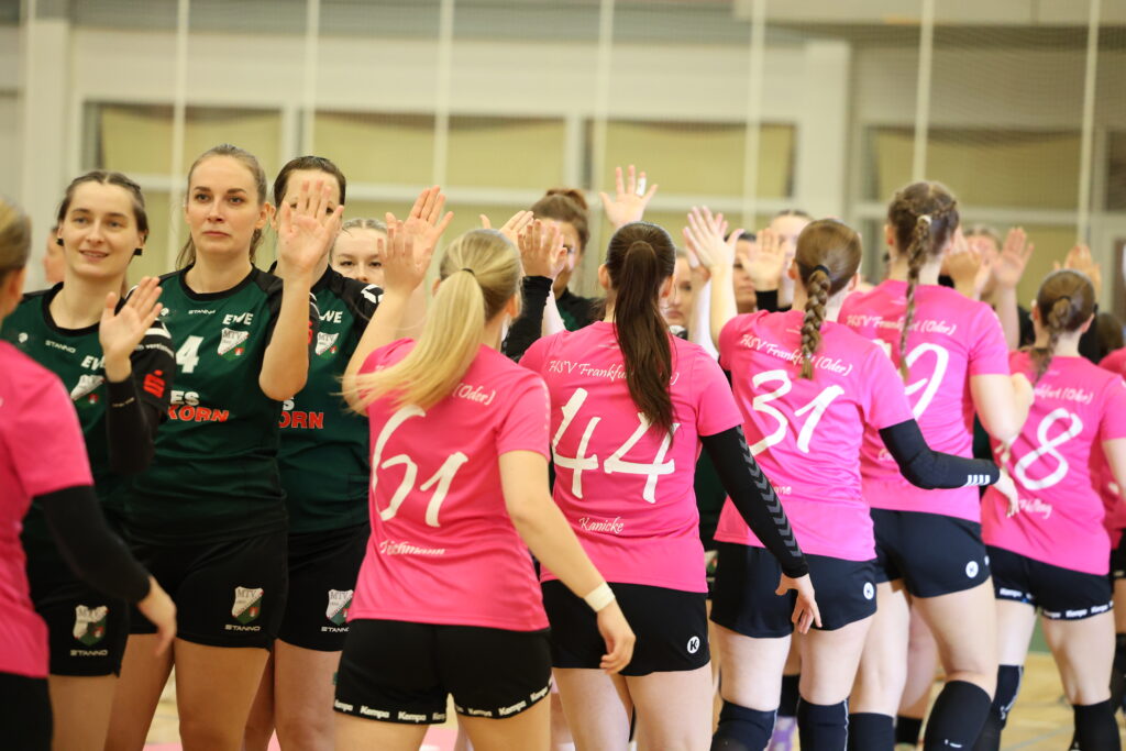 Zwei Frauenhandballmannschaften – eine in Grün und eine in Rosa – stellen sich auf dem Hallenfeld auf, um sich gegenseitig High Fives zu geben, und demonstrieren damit Teamgeist und Nachwuchsleistungssport, während ihre Trikots mit den Rückennummern von hinten zu sehen sind.