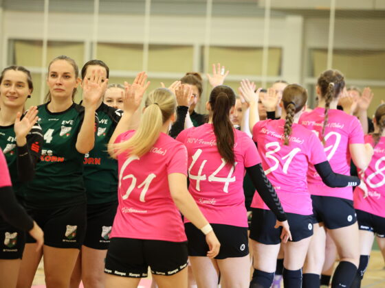 Zwei Frauenhandballmannschaften – eine in Grün und eine in Rosa – stellen sich auf dem Hallenfeld auf, um sich gegenseitig High Fives zu geben, und demonstrieren damit Teamgeist und Nachwuchsleistungssport, während ihre Trikots mit den Rückennummern von hinten zu sehen sind.