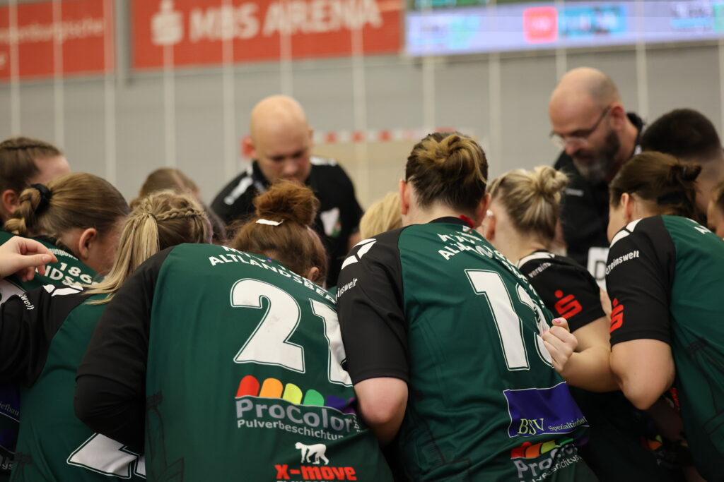 Eine Gruppe von Sportlern in grünen Trikots mit den Nummern 21 und 14 steht mit ihren Trainern in einer Sporthalle zusammen und bereitet sich auf ein Spiel im Nachwuchsleistungssport vor. Im Hintergrund sind eine unscharfe Anzeigetafel und das Schild „MIS ARENA“ zu sehen.