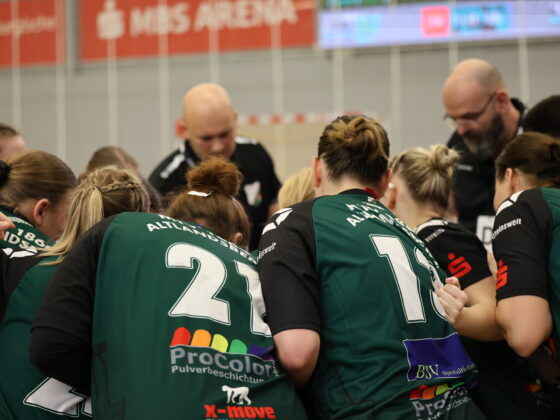 Eine Gruppe von Sportlern in grünen Trikots mit den Nummern 21 und 14 steht mit ihren Trainern in einer Sporthalle zusammen und bereitet sich auf ein Spiel im Nachwuchsleistungssport vor. Im Hintergrund sind eine unscharfe Anzeigetafel und das Schild „MIS ARENA“ zu sehen.