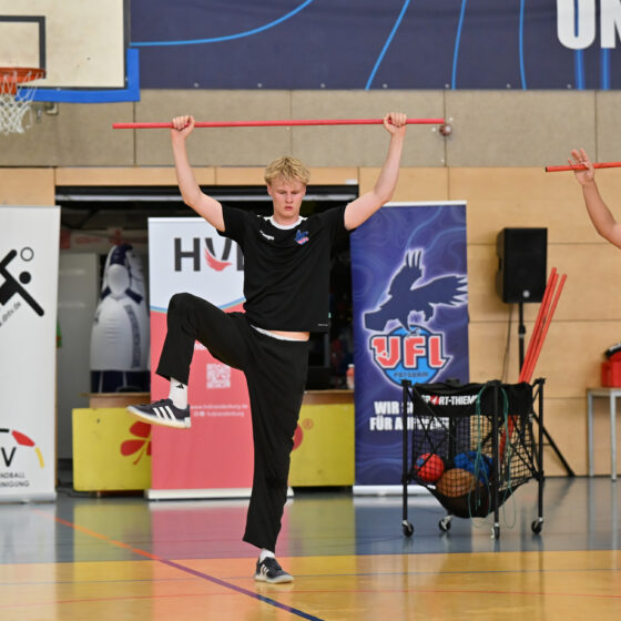 Zwei junge Männer in Sportkleidung balancieren in einer Sporthalle auf einem Bein und halten rote Stäbe über ihren Köpfen. Der Nachwuchsleistungssport wird durch Sportbanner, einen Basketballkorb und die im Hintergrund sichtbare Ausrüstung deutlich.