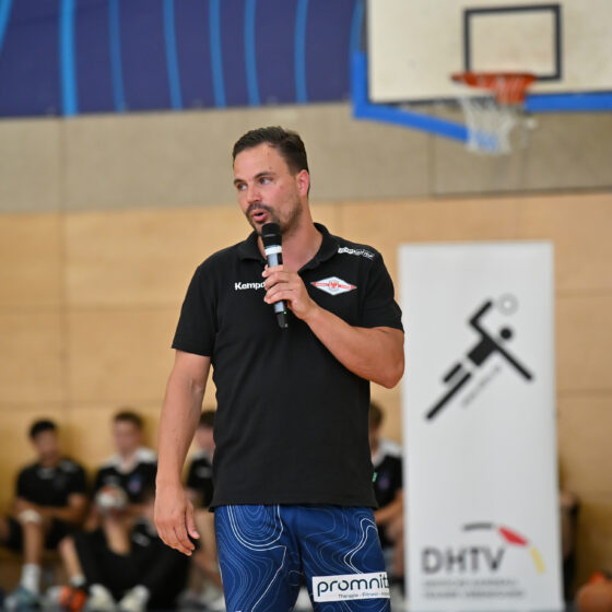 Ein Mann in einem schwarzen Hemd und blauen Shorts spricht in einer Sporthalle in ein Mikrofon und geht dabei vermutlich auf Themen im Zusammenhang mit dem Nachwuchsleistungssport ein. Hinter ihm sitzen Menschen, und man sieht Sportbanner, einen Basketballkorb und eine Holzwand.