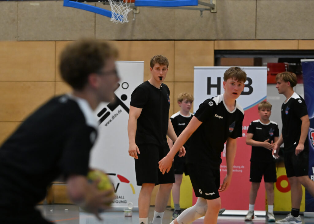Eine Gruppe junger männlicher Sportler in schwarzen Sporttrikots bestreitet ein Handballspiel im Rahmen des Nachwuchsleistungssports in der Halle; ein Spieler hält dabei den Ball in der Hand. Im Hintergrund beobachtet ein Schiedsrichter in schwarzer Kleidung das Geschehen aufmerksam und pfeift. Im Bild sind Banner und die Wände der Sporthalle zu sehen.