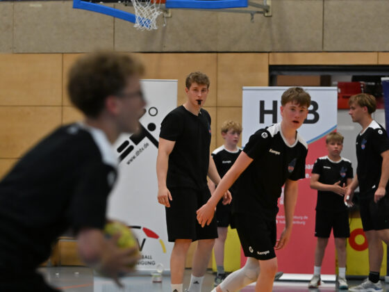 Eine Gruppe junger männlicher Sportler in schwarzen Sporttrikots bestreitet ein Handballspiel im Rahmen des Nachwuchsleistungssports in der Halle; ein Spieler hält dabei den Ball in der Hand. Im Hintergrund beobachtet ein Schiedsrichter in schwarzer Kleidung das Geschehen aufmerksam und pfeift. Im Bild sind Banner und die Wände der Sporthalle zu sehen.