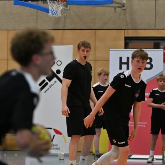 Eine Gruppe junger männlicher Sportler in schwarzen Sporttrikots bestreitet ein Handballspiel im Rahmen des Nachwuchsleistungssports in der Halle; ein Spieler hält dabei den Ball in der Hand. Im Hintergrund beobachtet ein Schiedsrichter in schwarzer Kleidung das Geschehen aufmerksam und pfeift. Im Bild sind Banner und die Wände der Sporthalle zu sehen.