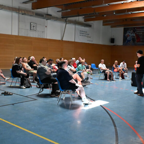 Eine Gruppe von Menschen sitzt auf Stühlen in einer Sporthalle und hört einem Redner zu, der vor ihnen steht. Die Sporthalle mit ihren Holzwänden, freiliegenden Balken und einem Handballtor bildet den Rahmen für Diskussionen über den Nachwuchsleistungssport.