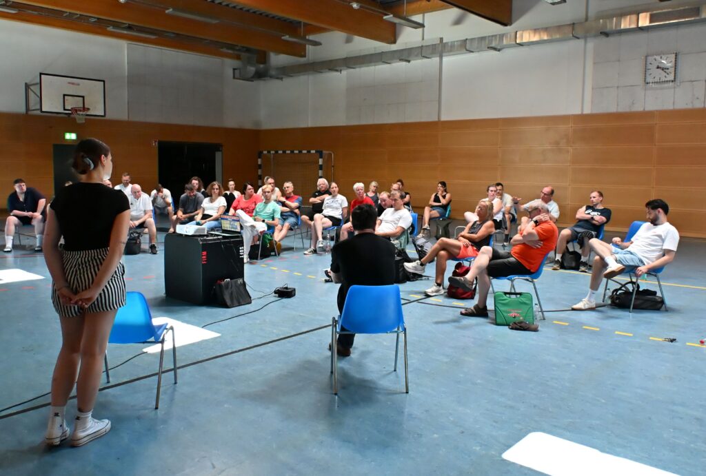 Eine Gruppe von Menschen sitzt auf Stühlen, die in einer Turnhalle aufgestellt sind, und hört einer Person zu, die vorne steht. Die Turnhalle, die für den Nachwuchsleistungssport genutzt wird, verfügt über Holzwände, einen Basketballkorb und eine Uhr an der Wand.