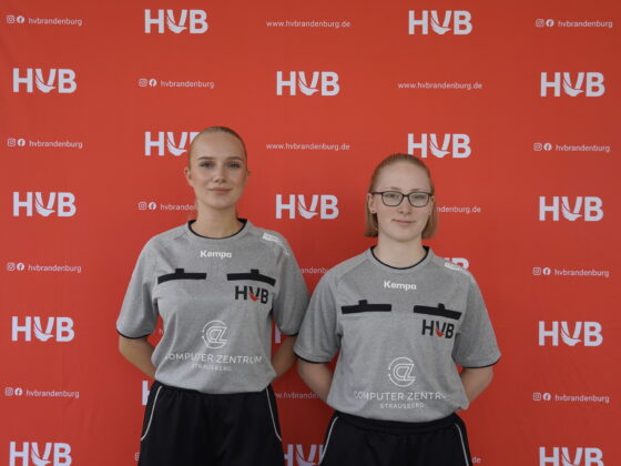 Zwei Frauen in grauen Sportshirts posieren vor einem roten Hintergrund, auf dem sich HVB-Logos und Website-Adressen wiederholen.
