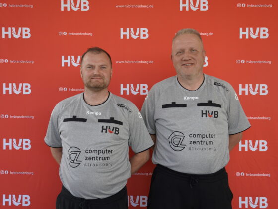 Zwei Männer in grauen Sporttrikots mit HVB-Logos stehen vor einem roten Hintergrund, auf dem sich der Schriftzug „HVB“ und das HVB-Logo wiederholen. Beide lächeln und blicken in die Kamera.