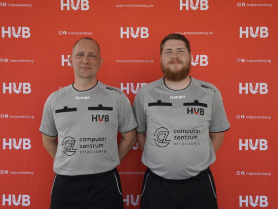 Zwei Männer in identischen grauen Sporthemden stehen lächelnd nebeneinander vor einem roten Hintergrund, auf dem wiederholt die Logos von HVB und der Website aufgedruckt sind.