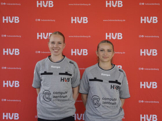 Zwei Frauen in grauen Sporttrikots stehen lächelnd nebeneinander vor einem roten Hintergrund, auf dem sich „HVB“-Logos und Website-Texte wiederholen.