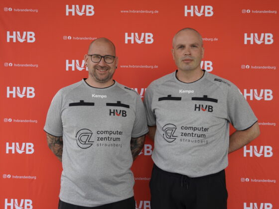 Zwei Männer stehen lächelnd nebeneinander und tragen identische graue Sportshirts mit der Aufschrift „HVB“ und Sponsorenlogos vor einem roten Hintergrund, auf dem sich „HVB“-Logos wiederholen.
