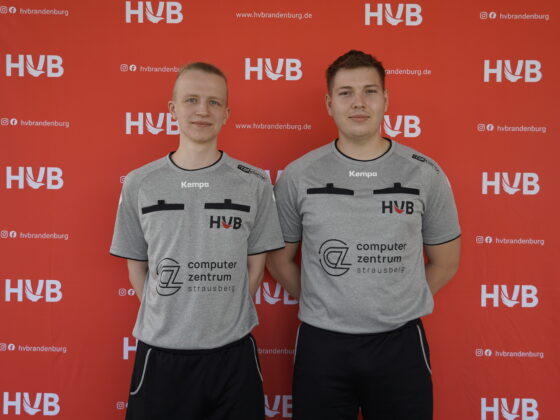 Zwei junge Männer in identischen grauen Sporthemden stehen nebeneinander vor einem roten Hintergrund, auf dem wiederholt die Buchstaben „HVB“ und Social-Media-Symbole aufgedruckt sind.