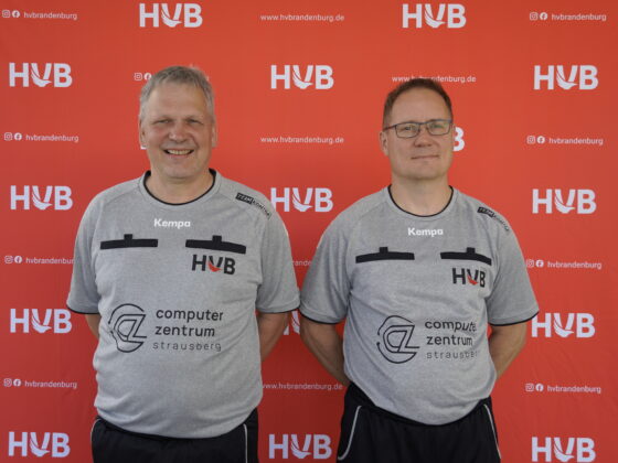 Zwei Männer in identischen grauen Sporthemden stehen nebeneinander und lächeln in die Kamera, während sie vor einem roten Hintergrund mit weißen HVB-Logos und Website-Text posieren.