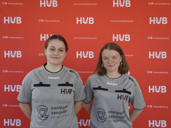 Zwei Frauen in grauen Shirts mit schwarzen Logos stehen vor einem leuchtend roten Hintergrund mit dem HVB-Logo und lächeln in die Kamera.