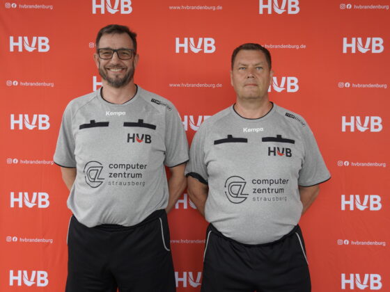 Zwei Männer in grauen Sporthemden und schwarzen Shorts stehen vor einem roten Hintergrund mit „HVB“-Logos. Der eine trägt eine Brille und lächelt, der andere blickt ernst. Beide haben die Hände hinter dem Rücken und Sponsorenlogos auf ihren Hemden.
