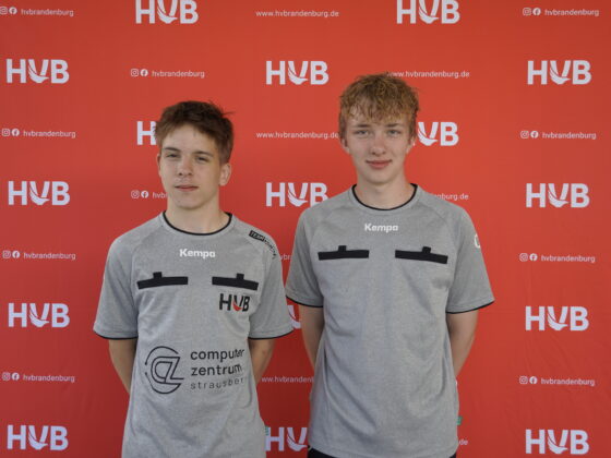 Zwei junge Leute in grauen Sportshirts stehen nebeneinander und posieren vor einem roten Hintergrund mit mehreren weißen HVB-Logos und Social-Media-Symbolen.