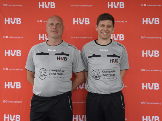 Zwei Männer in grauen Sporthemden mit HVBr-Logos stehen lächelnd vor einem roten Hintergrund, auf dem das HVB-Logo wiederholt zu sehen ist. Beide haben kurze Haare und tragen schwarze Shorts. Auf dem Hintergrund sind Website-Informationen und Social-Media-Symbole zu sehen.