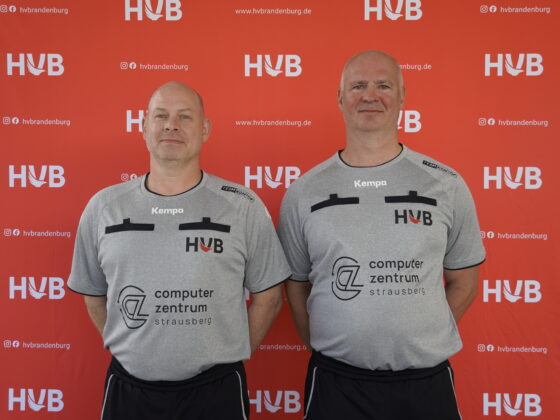 Zwei Männer, die identische graue Sporthemden und schwarze Shorts tragen, stehen lächelnd nebeneinander. Hinter ihnen befindet sich ein roter Hintergrund, auf dem wiederholt weiße HVB-Logos und Website-URLs aufgedruckt sind.