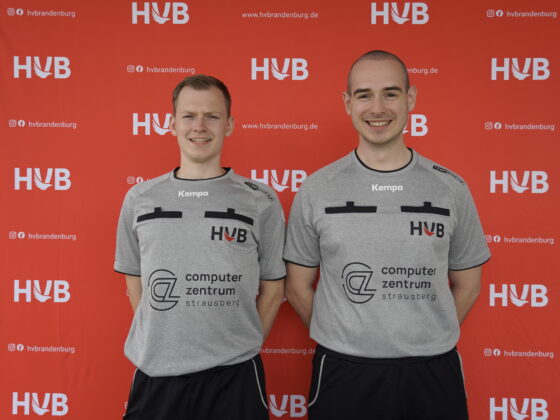 Zwei lächelnde Männer in identischen grauen Kempa-Schiedsrichtertrikots stehen vor einem roten Hintergrund, auf dem „HVB“ und andere Logos mehrfach aufgedruckt sind.