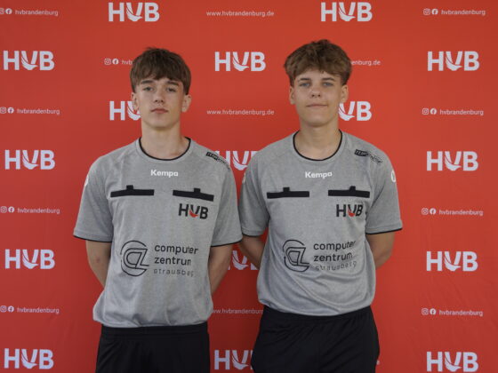 Zwei junge Männer in identischen grauen Sporthemden stehen nebeneinander und blicken nach vorne, vor einem roten Hintergrund mit weißen HVB- und Website-Logos.