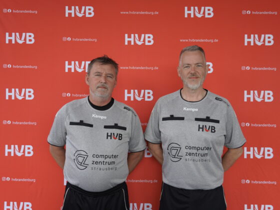 Zwei Männer in identischen grauen Sporthemden stehen vor einem roten Hintergrund mit HVB-Logos und Website-Text und blicken mit neutralen Gesichtsausdrücken in die Kamera.