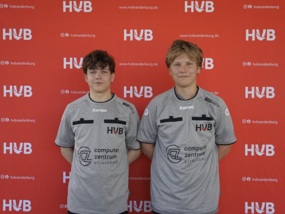 Zwei Teenager in identischen grauen Sporttrikots stehen vor einem roten Hintergrund mit weißen HVB-Logos und Schriftzügen. Beide haben kurzes, dunkelblondes oder braunes Haar und lächeln leicht in die Kamera.
