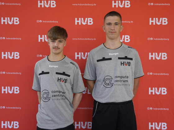 Zwei junge Männer in identischen grauen Sporthemden stehen vor einem roten Hintergrund, auf dem sich HVB-Logos und Website-Texte wiederholen. Beide blicken in die Kamera und scheinen zum selben Team zu gehören.