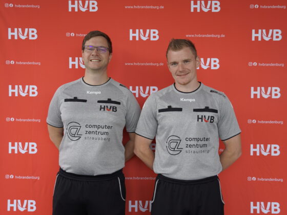 Zwei Männer in identischen grauen Sporthemden stehen lächelnd vor einem roten Hintergrund, auf dem sich „HVB“-Logos und Schriftzüge wiederholen. Beide haben die Hände hinter dem Rücken.