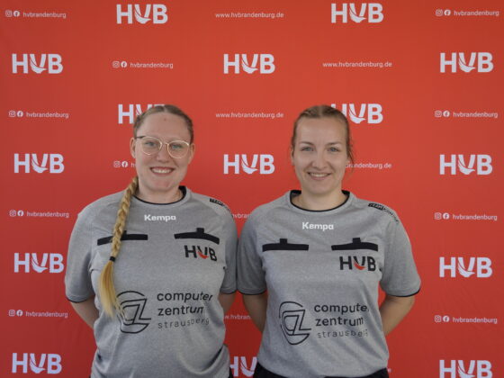 Zwei Frauen in identischen grauen Sporttrikots stehen lächelnd vor einem roten Hintergrund mit weißen HVB-Logos und Social-Media-Handles.