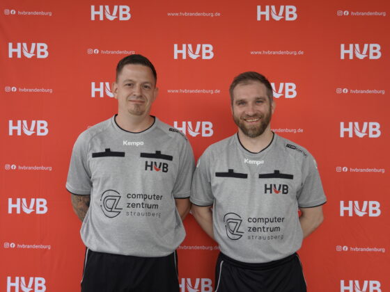Zwei Männer in grauen Sporttrikots stehen nebeneinander und lächeln vor einem roten Hintergrund mit dem HVB-Logo. Die Trikots sind mit Sponsorenlogos versehen und weisen schwarze Akzente an den Schultern und auf der Brust auf.