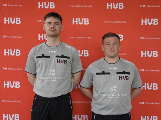 Zwei Männer in grauen Sporttrikots stehen nebeneinander vor einem roten Hintergrund mit dem HVB-Logo und anderen Logos. Beide blicken nach vorne und posieren für ein Mannschaftsfoto.