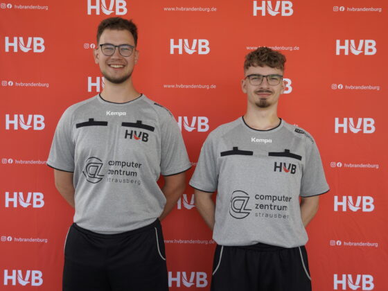 Zwei Männer in grauen Sporthemden mit Logos stehen lächelnd nebeneinander vor einem roten Hintergrund, auf dem wiederholt „HVB“ und der Text der Website aufgedruckt sind.