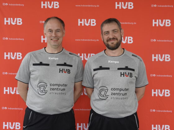 Zwei Männer in grauen Sporthemden mit schwarzem Besatz stehen lächelnd vor einem roten Hintergrund mit weißen HVB-Logos und Schriftzügen. Auf den Hemden sind Logos des „Computerzentrums Strausberg“ sowie das HVB-Logo zu sehen.