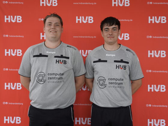 Zwei Männer in grauen Sporttrikots stehen nebeneinander vor einem roten Hintergrund mit den Logos von HVB und hvbrandenburg.de. Beide lächeln und blicken in die Kamera.
