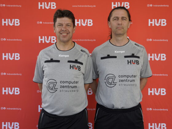 Zwei Männer in grauen Sporthemden stehen lächelnd nebeneinander vor einem roten Hintergrund mit weißen „HVB“-Logos und Social-Media-Symbolen.