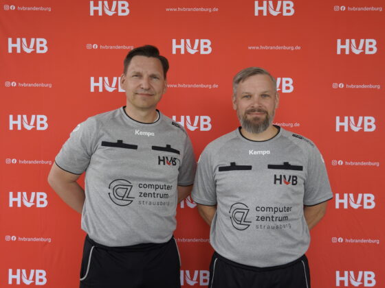 Zwei Männer in identischen grauen Hemden mit HVB-Logos stehen lächelnd nebeneinander vor einem roten Hintergrund mit dem HVB-Logo.