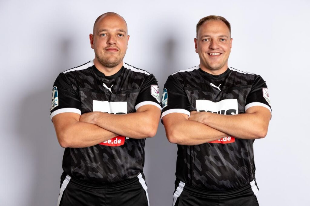 Zwei Männer mit kurzen Haaren, die identische schwarze Sporttrikots mit weißen Details tragen, stehen Seite an Seite mit verschränkten Armen, blicken nach vorne und lächeln vor einem schlichten, hellen Hintergrund – ein selbstbewusstes Duo, das den Geist des Nachwuchsleistungssports verkörpert.