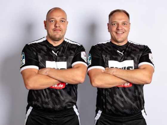 Zwei Männer mit kurzen Haaren, die identische schwarze Sporttrikots mit weißen Details tragen, stehen Seite an Seite mit verschränkten Armen, blicken nach vorne und lächeln vor einem schlichten, hellen Hintergrund – ein selbstbewusstes Duo, das den Geist des Nachwuchsleistungssports verkörpert.