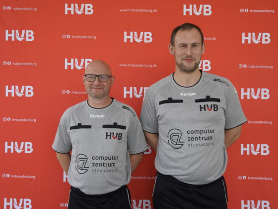 Zwei Männer in grauen Sporthemden mit HVB- und Sponsorenlogos stehen lächelnd vor einem roten Hintergrund, auf dem sich der Schriftzug „HVB“ und der Website-Text wiederholen.