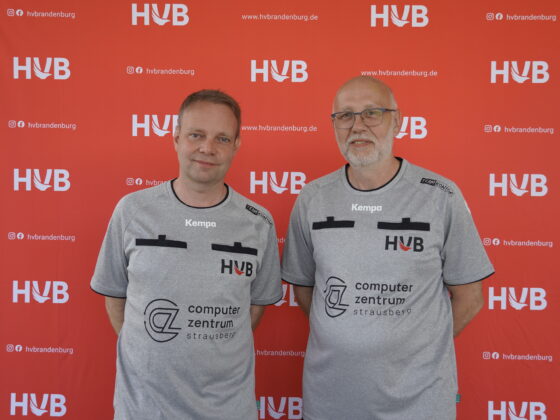 Zwei Männer in identischen grauen Sporthemden stehen vor einem roten Hintergrund mit dem HVB-Logo. Beide lächeln sanft, und auf den Hemden sind das Kempa-Logo sowie der Schriftzug „Computerzentrum Strausberg“ zu sehen.