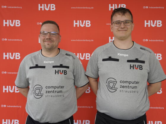 Zwei Männer in grauen Sporthemden mit den Aufdrucken „HVB“ und „Computer Zentrum Strausberg“ stehen vor einem roten Hintergrund mit weißen HVB-Logos. Beide lächeln und blicken in die Kamera.