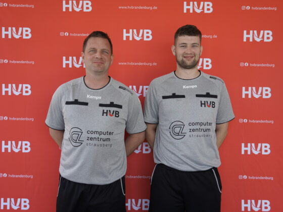 Zwei Männer in grauen HVB-Shirts stehen lächelnd nebeneinander vor einem roten Hintergrund, auf dem „HVB“ und Social-Media-Symbole aufgedruckt sind.