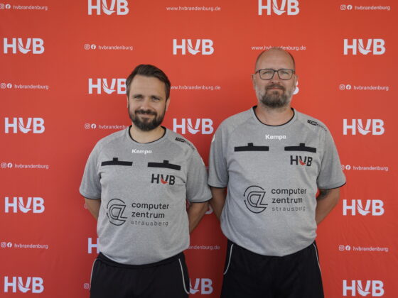 Zwei Männer in grauen Sporthemden stehen nebeneinander vor einem roten Hintergrund mit mehreren HVB-Logos. Beide tragen eine Brille und haben Bärte. Die Hemden sind mit schwarzen Akzenten und verschiedenen Sponsorenlogos versehen.