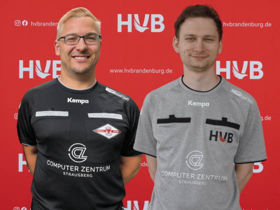 Zwei Männer stehen lächelnd vor einem roten Hintergrund mit weißen „HVB“-Logos. Beide tragen sportliche Shirts mit Logos und Abzeichen – typisch für die Funktionäre von Schiedsrichter Handball; der Mann links ist schwarz gekleidet, während der Mann rechts Grau trägt.