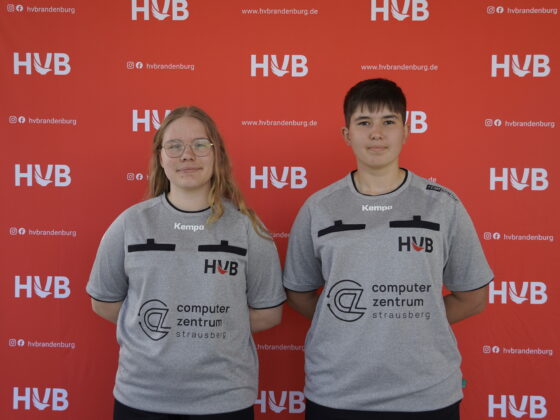 Zwei Personen in grauen Team-Shirts stehen vor einem roten Hintergrund, auf dem sich die Logos von HVB und der Website wiederholen. Beide blicken in die Kamera, stehen nebeneinander und haben die Hände hinter dem Rücken.
